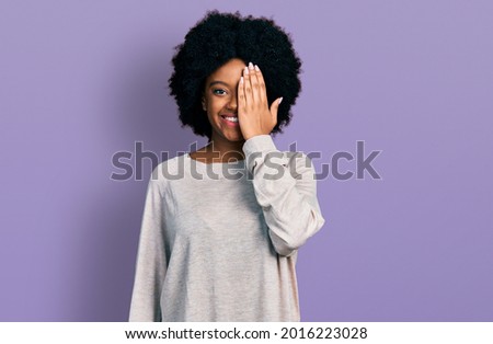 Young african american woman wearing casual clothes covering one eye with hand, confident smile on face and surprise emotion. 
