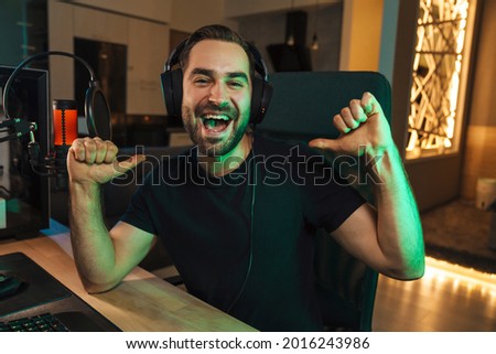 Happy young man gamer streamer in headphones playing on computer talking with players on chat in gaming competition celebrating win