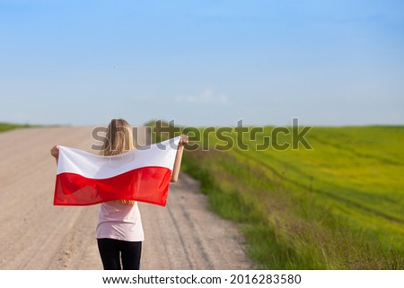 Blond girl holding flag of Poland walking down unpaved road. Back view. Polish Flag Day. Independence Day. Travel and learn polish language concept. Royalty-Free Stock Photo #2016283580