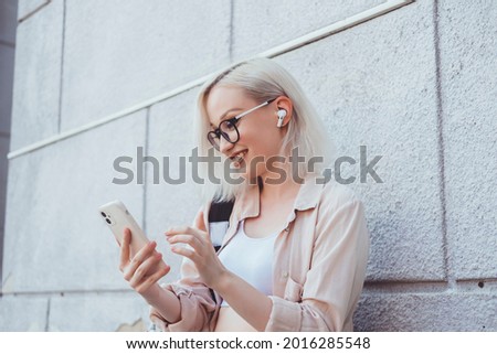 Young blonde woman using smartphone in hand, outdoor hipster portrait, street style, glasses, happy face, backpack, earphones, jacket Royalty-Free Stock Photo #2016285548