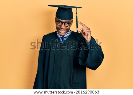Young african american man wearing graduation cap and ceremony robe smiling and confident gesturing with hand doing small size sign with fingers looking and the camera. measure concept. 