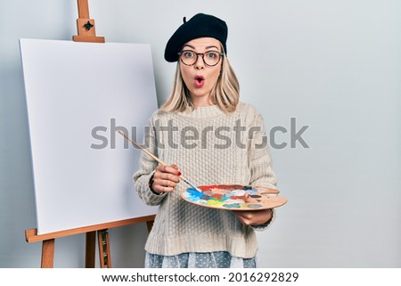 Beautiful caucasian woman drawing with palette on easel stand afraid and shocked with surprise and amazed expression, fear and excited face. 