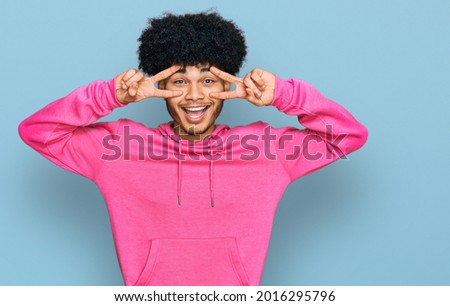 Young african american man with afro hair wearing casual pink sweatshirt doing peace symbol with fingers over face, smiling cheerful showing victory 