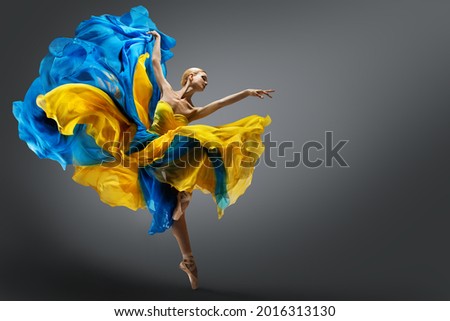 Beautiful Woman Ballet Dancer Jumping in Air in Colorful Fluttering Dress. Graceful Ballerina Dancing in Yellow Blue Gown over Gray Studio Background Royalty-Free Stock Photo #2016313130