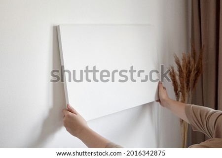 Blank canvas in female hands, picture mockup. Woman hanging canvas on white wall in interior with dry grass and beige curtains