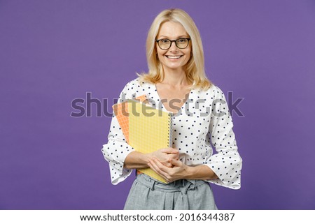 Smiling gray-haired blonde teacher woman lady 40s 50s years old wearing white dotted blouse eyeglasses standing hold notepads looking camera isolated on bright violet color background studio portrait