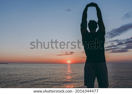 Silhouette rear view young strong sporty athletic toned fit sportsman man 20s wearing sports clothes warm up training at sunrise sun dawn over sea beach outdoor on pier seaside in summer day morning.