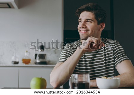 Young happy man in striped t-shirt eat breakfast muesli cereals with milk fruit in bowl drink coffee sit by table look aside cooking food in light kitchen at home alone Healthy diet lifestyle concept. Royalty-Free Stock Photo #2016344534