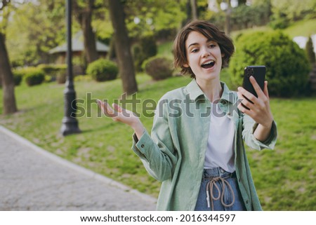 Young woman 20s in casual green jacket jeans walking strolling in city spring park outdoors, talk by video call mobile cell phone spread hand. People active urban healthy lifestyle cycling concept.