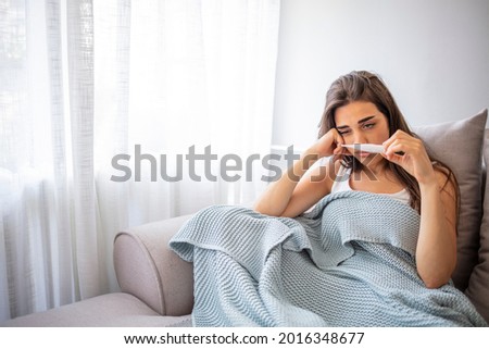 Woman with fever symptoms sitting on sofa and holding thermometer. One woman, young woman with Covid-19 symptoms sitting on sofa at home, holding thermometer.