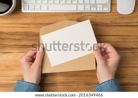 First person top view photo of hands holding open craft paper envelope and white card cup keyboard and mouse on isolated wooden table background with blank space