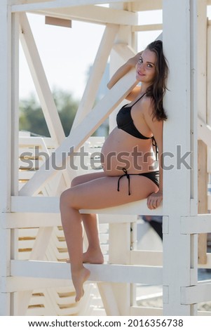 Expectant mother on the beach. Attractive pregnant woman in black bikini sitting with one arm raised on white boards of rescue booth on background of beach beds, looking at camera and smiling