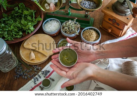 Women's hands holding a tin jar of homemade salve. Home herbalism and cosmetics. Natural homemade salve in metallic tin jar with dried plants and herbs flowers. Royalty-Free Stock Photo #2016402749
