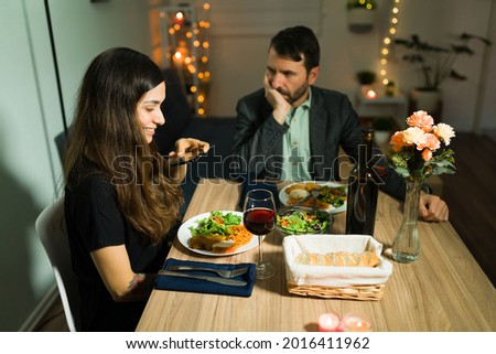 Excited young woman taking a picture of her delicious dinner during a fancy date at home with her bored upset boyfriend