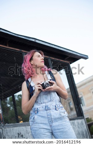 half-length woman casually dressed with pink hair holding a retro camera outdoors
