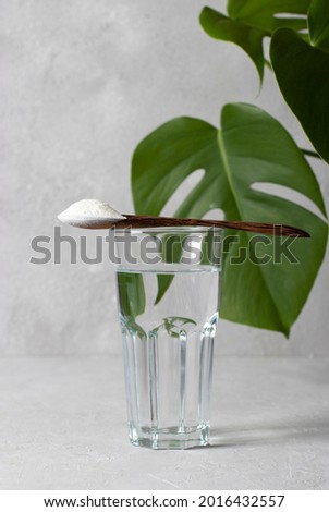 Collagen powder in spoon on glass of water on light gray background with monstera leaf. Healthy and anti age concept.