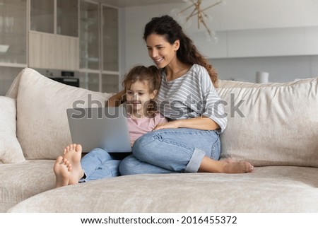 Happy young Hispanic mother and small biracial daughter relax on sofa in living room using computer together. Smiling Latino mom and ethnic girl child rest on couch talk on video call on laptop.