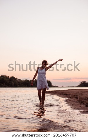 Photo of a dancing woman at sunset. Beautiful blonde in a white summer dress on the river bank against the background of the beach and the beach. 