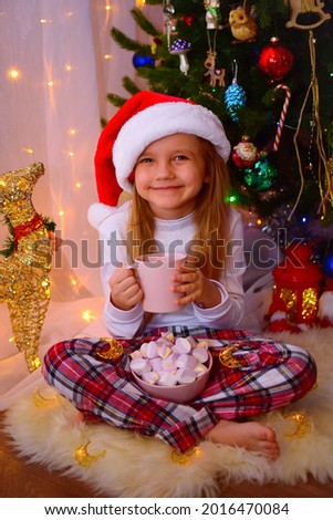 A little cute girl in a red santa claus hat sits on a white sheep's skin against the background of a Christmas tree with a mug of cocoa in her hands and a Minsk marshmallow and smiles sweetly