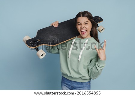 Positive, naughty girl holding a skate on her shoulder, showing a rock gesture. Teen girl isolated on blue background