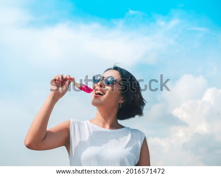 Smiling Asian woman short hair in casual white sleeveless shirt wearing sunglasses holding pink popsicle on blue sky background in summertime. Women eating popsicles, freshy summer season concept.