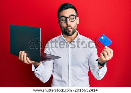 Young hispanic man wearing business style holding laptop and credit card making fish face with mouth and squinting eyes, crazy and comical. 