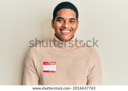 Young handsome hispanic man wearing hello my name is sticker identification looking positive and happy standing and smiling with a confident smile showing teeth 