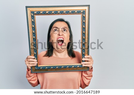 Middle age hispanic woman holding empty frame angry and mad screaming frustrated and furious, shouting with anger looking up. 