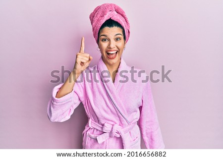 Young hispanic woman wearing shower towel cap and bathrobe pointing finger up with successful idea. exited and happy. number one. 