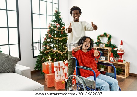 Young interracial couple with woman sitting on wheelchair by christmas tree approving doing positive gesture with hand, thumbs up smiling and happy for success. winner gesture. 