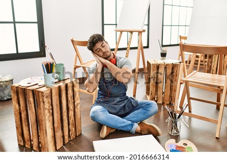 Young hispanic man sitting at art studio sleeping tired dreaming and posing with hands together while smiling with closed eyes. 