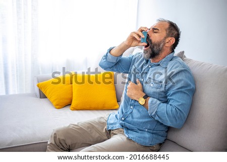 Cropped shot of a handsome mature man sitting alone at home and using an asthma pump. Man Inhaling Asthmatic Cure at Home. Mature man using medical inhaler to prevent shortness of breath Royalty-Free Stock Photo #2016688445