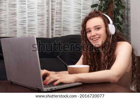 Caucasian teenage girl doing homework on her computer with headphones on while looking at you and smiling.