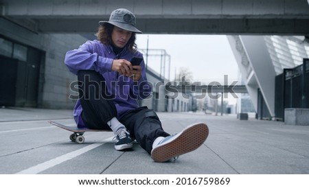 Young sporty man reading message online on phone on skateboard. Stylish hipster sitting on skate with mobile phone outdoor. Cool skater guy holding smartphone outside. Royalty-Free Stock Photo #2016759869