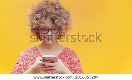 Attractive girl wearing nerdy spectacles holding a mobile phone in her hand. Typing a text on her cellphone and smiling. Concept of online text messages and social media. Bright yellow background