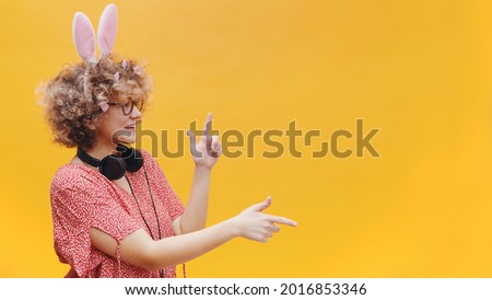 Attractive young girl wearing cute bunny ears and spectacles in a joyful mood. Headphones around her neck. Girl smiling and pointing at something. Bright yellow background. Studio shot.