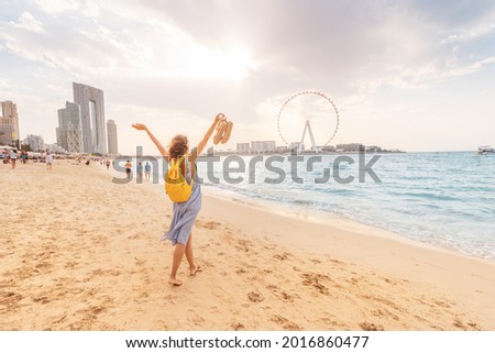 Happy female tourist walks barefoot on a sandy beach in the JBR area of Dubai and admires the panoramic view of the Ain Ferris Wheel Royalty-Free Stock Photo #2016860477