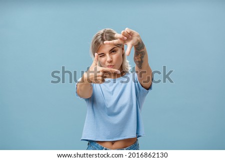 Artistic and creative female designer in stylish t-shirt closing one eye folding lip and making frame gesture while looking through it as if taking measurement or picturing something over blue wall