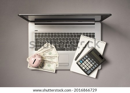 Top view photo of piggy bank on fan of money dollars calculator pen and copybook on open grey laptop on isolated grey background