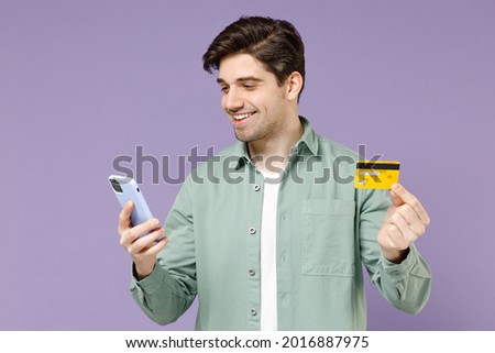 Young fun happy caucasian man 20s wear casual mint shirt white t-shirt hold mobile cell phone credit bank card shopping online isolated on purple background studio portrait People lifestyle concept