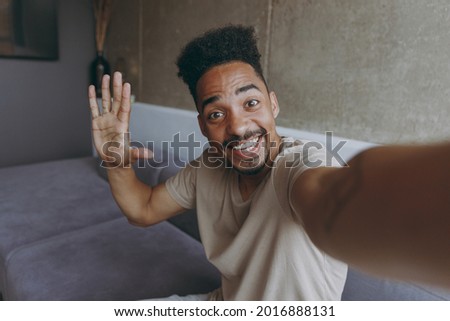 Close up fun happy smiling young african american man in beige casual t-shirt sit on grey sofa indoors apartment doing selfie shot on mobile phone waving hand say hello, rest on weekends stay at home.