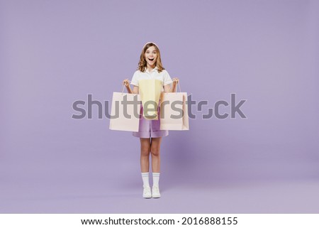 Full length excited surprised little kid girl 12-13 years old wear white shirt holding package bags with purchases after shopping isolated on purple background. Childhood children lifestyle concept