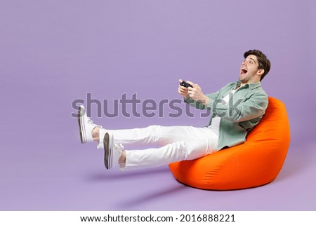 Full length man in casual mint shirt white t-shirt sitting in orange bean bag chair hold takeaway bucket eat popcorn watch movie film play pc game with joystick console isolated on purple background. Royalty-Free Stock Photo #2016888221