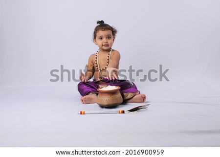 adorable Indian baby in krishna kanha or kanhaiya dress posing with his flute and dahi handi (pot with curd) on white background. sitting pose