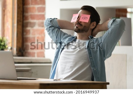Peaceful happy young man employee worker with colorful sticky paper notes on eyes relaxing on chair, enjoying break pause stress free time during lazy working day in modern office, deadline concept. Royalty-Free Stock Photo #2016949937