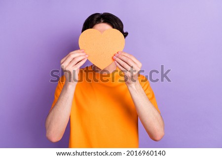Photo of young man hold hands orange paper heart shape cover face isolated on purple violet color background
