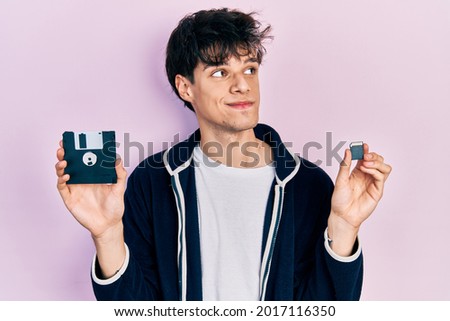Handsome hipster young man holding floppy disk and sdxc card smiling looking to the side and staring away thinking. 