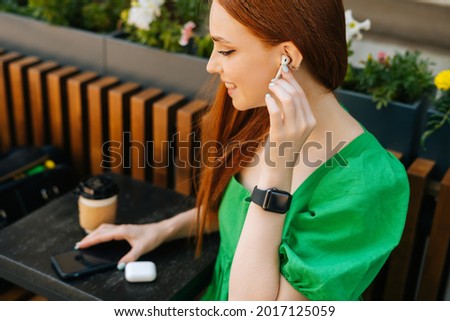 Close-up top view of smiling young woman touching wireless earphones, listening music using mobile phone sitting at table, in outdoor cafe terrace in summer day, blurred background, selective focus