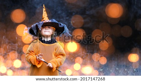 Happy Halloween concept.Cute funny little child with a magic wand on dark background with glowing bokeh lights.Beautiful girl in a witch orange costume conjures in the night. Banner with copy space