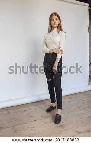 portrait of thoughtful teen model wearing white sweater and black jeans. caucasian skinny young female with blond hair stands in front of white paper background. natural pretty lady standing at studio